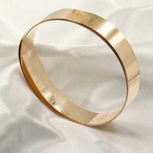 May include: A gold-coloured bangle bracelet with a flat, wide design. The bracelet is made of a shiny metal and is set against a white, textured background. The bangle is a simple, elegant piece of jewellery.