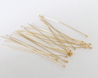 May include: A pile of gold-colored head pins with round heads. The pins are scattered on a white surface.