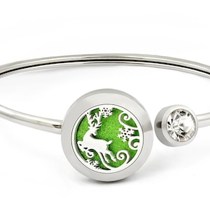 May include: A silver bangle bracelet with a round, green, and silver charm featuring a deer silhouette and snowflakes. The charm has a clear crystal on the opposite side.
