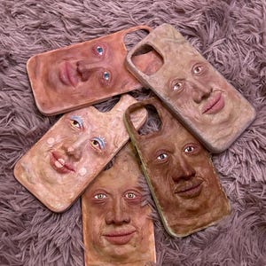 Op de afbeelding: Een verzameling van vijf unieke telefoonhoesjes, elk met een gebeeldhouwd menselijk gezicht. De hoesjes variëren in huidskleur, met gedetailleerde gelaatstrekken zoals ogen, lippen en neuzen. De hoesjes zijn gerangschikt op een zacht, getextureerd oppervlak.