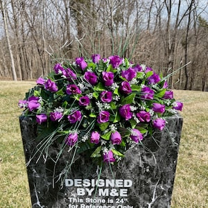 May include: A black marble-like headstone with a floral arrangement of purple roses and green foliage. The headstone has the text "DESIGNED BY M&E" and "This Stone is 24" for Reference Only".