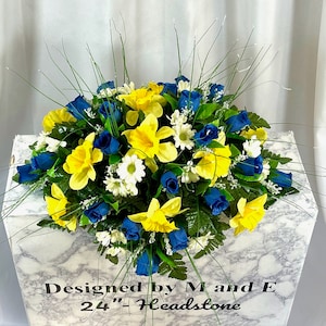 May include: A blue and yellow floral arrangement with white daisies, blue roses, and yellow daffodils. The arrangement is on a white marble headstone with the text "Designed by M and E 24" Headstone".