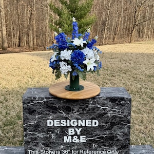 May include: A floral arrangement featuring blue, white, and green artificial flowers. The flowers are in a green vase, sitting on a wooden base. The base rests on a black marble-like stone. Text on the stone reads "DESIGNED BY M&E".