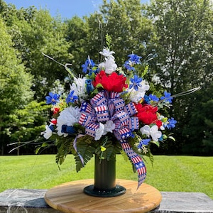 May include: A patriotic floral arrangement featuring red, white, and blue flowers, including roses and daisies. A large bow with a stars and stripes pattern is the centerpiece. The arrangement is in a dark green vase, set on a wooden spool.