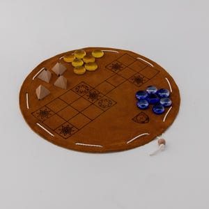 Peut inclure: Un jeu de société en cuir marron avec un bord cousu, un motif quadrillé, des pyramides en bois et des pièces de jeu en verre jaunes et bleues. Une petite perle en bois est attachée au plateau avec une ficelle.