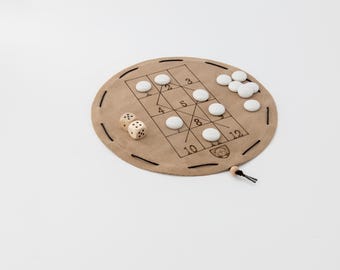 May include: A circular tan leather game board featuring a printed grid with numbers 2 through 12, black stitched edges, and a drawstring closure. The set includes two wooden dice with black pips and several smooth white circular game tokens.