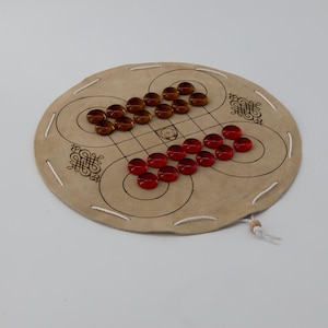 Könnte beinhalten: Ein rundes, braunes Leder-Spielbrett mit einem weißen Schnurbord. Das Brett hat ein Muster aus Kreisen und Linien, und es gibt rote und braune Glas-Spielsteine auf dem Brett.
