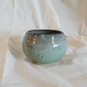 May include: A small, rounded ceramic bowl featuring a smooth, glossy glaze that transitions from a deep, dark blue at the rim to a lighter, muted sage green at the base. The surface displays subtle, speckled texture throughout the finish.