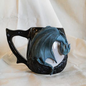 Peut inclure: Une tasse en céramique noire ornée d'un dragon bleu sculpté en trois dimensions sur le côté. La tasse possède une anse noire brillante et une base texturée, avec les ailes et la tête du dragon qui ressortent de la surface lisse.