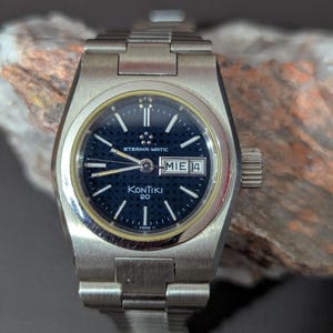 May include: A silver-toned Eterna-Matic Kontiki 20 wristwatch with a dark blue face. The watch has a date display and a silver band. The watch face has silver hands and markers. The watch is Swiss made.