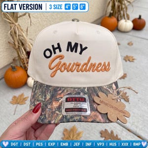 May include: A cream-colored baseball cap with a camouflage-patterned brim. The front features the embroidered text "OH MY" in black and "Gourdness" in orange. A sticker on the brim reads "OTTO CLASSIC FIT OSFM," and a flower-shaped tag says "MINOSHStudio."