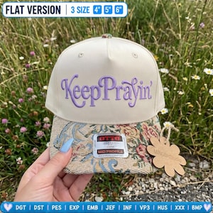 Könnte beinhalten: Eine cremefarbene Baseballkappe mit dem violett gestickten Schriftzug KeepPrayin' auf der Vorderseite. Der Schirm ist mit einem mehrfarbigen floralen Gobelinstoff bezogen. Ein Aufkleber auf dem Schirm zeigt die Aufschrift Otto Classic Fit.