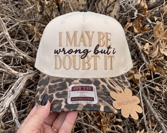 Puede incluir: Una gorra de béisbol color crema con visera de estampado de leopardo. La parte frontal presenta el texto bordado I MAY BE wrong but i DOUBT IT. La gorra incluye una pegatina en la visera y una etiqueta con forma de flor.