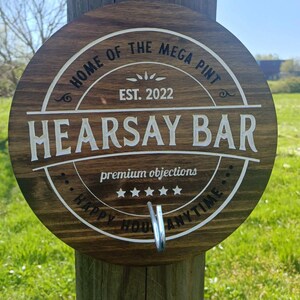 May include: A brown wooden sign with the text "Home of the Mega Pint, Est. 2022, Hearsay Bar, Premium Objections, Happy Hour Anytime". The sign has a metal hook attached to the bottom.