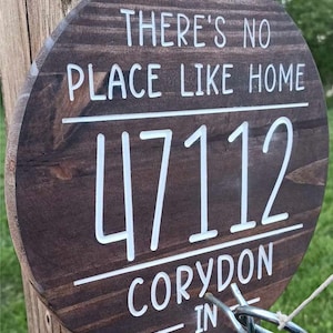 May include: A brown wooden sign with white text that reads "There's no place like home 47112 Corydon - IN". The sign is attached to a wooden post with a metal ring.
