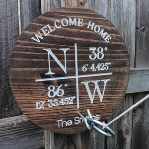 May include: A wooden round sign with the text "Welcome Home" and coordinates "N 38° 6' 4.425" and "W 86° 12' 35.452" and the text "The Smiths". The sign is attached to a wooden fence post with a metal ring.