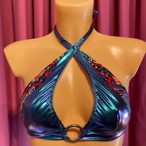Puede incluir: Un top halter azul iridiscente con estampado floral colorido y detalle de anillo plateado. El top tiene una abertura de ojo de cerradura en la parte delantera.