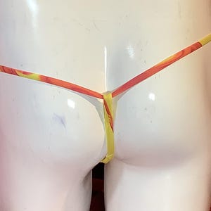 K&ouml;nnte beinhalten: Ein Tanga mit einem leuchtend gelben und orangefarbenen Marmormuster. Das Kleidungsst&uuml;ck ist mit d&uuml;nnen, minimalistischen Riemen konstruiert, die in der hinteren Mitte zusammenlaufen und ein schlichtes Design f&uuml;r diese Unterw&auml;sche bilden.