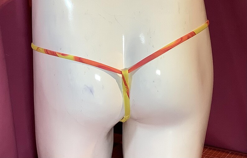 K&ouml;nnte beinhalten: Ein Tanga mit einem leuchtend gelben und orangefarbenen Marmormuster. Das Kleidungsst&uuml;ck ist mit d&uuml;nnen, minimalistischen Riemen konstruiert, die in der hinteren Mitte zusammenlaufen und ein schlichtes Design f&uuml;r diese Unterw&auml;sche bilden.