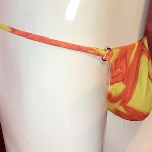 Può includere: Un perizoma caratterizzato da un vivace motivo marmorizzato giallo e arancione. Il capo è realizzato con sottili laccetti laterali che si collegano alla tasca anteriore tramite piccoli anelli metallici circolari, creando un netto contrasto.