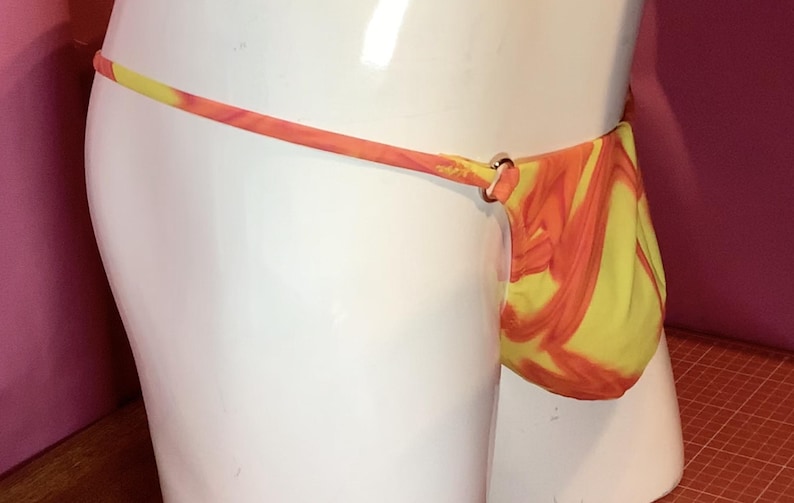 K&ouml;nnte beinhalten: Ein Tanga mit einem leuchtend gelben und orangefarbenen Marmormuster. Das Kleidungsst&uuml;ck ist mit d&uuml;nnen Seitenriemen konstruiert, die mit kleinen, kreisf&ouml;rmigen Metallringen an der Vordertasche befestigt sind und einen deutlichen Kontrast bilden.