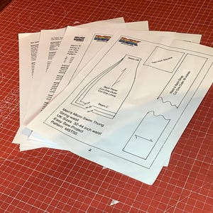 K&ouml;nnte beinhalten: Ein Stapel wei&szlig;er Papierb&ouml;gen mit einem Schnittmuster f&uuml;r einen Micro-Bade-String. Das oberste Blatt zeigt technische Strichzeichnungen, ein 5-cm-Testquadrat sowie Text, darunter Micro Swim Thong, UK-Gr&ouml;&szlig;en 32-44 und Pattern MST50.