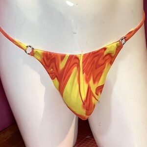 K&ouml;nnte beinhalten: Ein Tanga mit einem lebhaften gelb-orangen Marmormuster. Das Kleidungsst&uuml;ck ist mit d&uuml;nnen Seitenb&auml;ndern konstruiert, die an beiden Seiten durch kleine, kreisf&ouml;rmige silberfarbene Metallringe mit dem Vorderteil verbunden sind.
