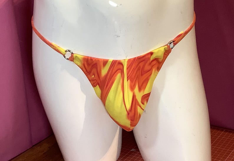 K&ouml;nnte beinhalten: Ein Tanga mit einem lebhaften gelb-orangen Marmormuster. Das Kleidungsst&uuml;ck ist mit d&uuml;nnen Seitenb&auml;ndern konstruiert, die an beiden Seiten durch kleine, kreisf&ouml;rmige silberfarbene Metallringe mit dem Vorderteil verbunden sind.