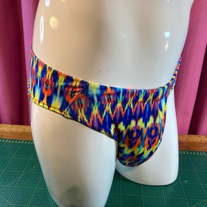 Può includere: Un paio di slip da bagno colorati con un motivo astratto blu, giallo, arancione e rosso. Gli slip sono realizzati in tessuto elastico e hanno un'altezza bassa.