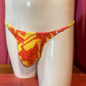 Puede incluir: Un tanga de traje de baño con estampado amarillo y naranja. La tela tiene un diseño arremolinado. El traje de baño se exhibe en un maniquí blanco. Las correas son finas y ajustables.
