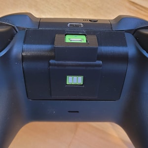 Puede incluir: Un mando de videojuegos negro con un puerto de carga verde y un indicador de batería que muestra una carga completa.