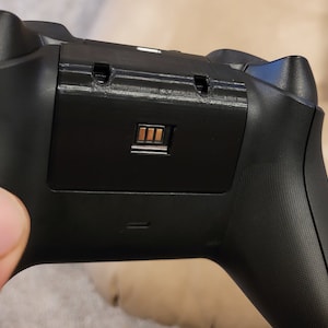 Puede incluir: Primer plano de un mando de videojuego negro con un pequeño conector rectangular dorado en la parte posterior.