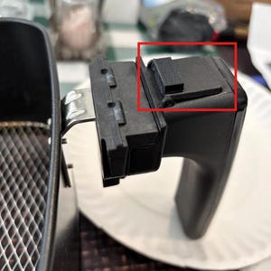 May include: Black air fryer handle with a small, rectangular, 3D-printed component attached. The handle is part of a larger appliance, with a metal basket visible on the left. The handle is on a white paper plate.