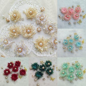 Puede incluir: Surtido de adornos florales de tela hechos a mano en varios colores, incluyendo blanco, amarillo, rosa, azul, rojo y verde. Cada flor presenta un centro de perla y detalles dorados. Algunos arreglos incluyen pequeñas perlas y dijes dorados de mariposa o nota musical. La palabra "INVALI" es visible.