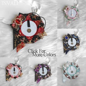 May include: A collection of decorative brooches in various colors. Each brooch features a central white circle with a black vertical stripe, surrounded by fabric and embellishments. The brooches have a ribbon and a pin on the back. Text on the image reads "Click For More Colors."