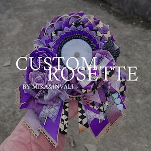 Könnte beinhalten: Eine individuelle Rosette in Lilatönen, mit einem zentralen weißen Kreis und dekorativen Bändern. Die Rosette enthält eine Stoffrose, goldene Akzente und den Text "CUSTOM ROSETTE" und "BY MIKA&INVALI".