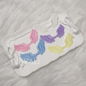 Peut inclure: Un plateau rectangulaire blanc avec des détails de nœuds décoratifs présente six ornements en forme d'ailes colorées. Les ailes sont dans des tons de rose, bleu, violet, jaune et blanc. Le mot "INVALI" est imprimé sur le plateau.