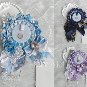 May include: Three decorative rosette brooches in blue, navy, and purple. Each brooch features layered ribbons, fabric rosettes, and a central felt circle. Embellishments include decorative brooches and ribbons with printed patterns. The brooches are displayed on a white surface.