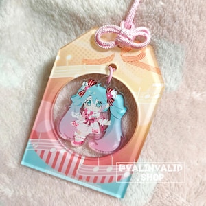 Peut inclure: Un charme en acrylique coloré représentant un personnage de dessin animé avec des cheveux bleus et une robe rose. Le charme est attaché à une ficelle rose et pend à une étiquette en acrylique transparente avec un motif de note de musique. L'étiquette porte le texte "#VALINVALID SHOP" imprimé dessus.
