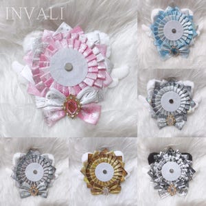 Puede incluir: Una colección de seis insignias circulares tipo roseta con cinta plisada en varios colores, incluyendo rosa, azul, plata, oro y negro. Cada insignia tiene un círculo central de fieltro, un lazo decorativo con una piedra preciosa y alas de tela blanca.