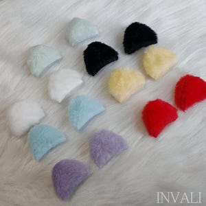 May include: A collection of small, fluffy, half-circle shaped objects in various colors, including white, black, yellow, red, blue, and purple. The objects are arranged on a white, textured surface. The word "INVALI" is visible in the bottom right corner.