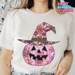 May include: White t-shirt featuring a pink pumpkin with a witch hat design. The pumpkin and hat are decorated with floral patterns in shades of pink, purple, and orange. The shirt has the text "ARTSY DIGITAL STUDIOS" in a colorful font.