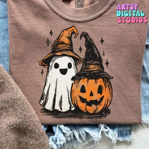 May include: A brown Comfort Colors sweatshirt featuring a Halloween-themed graphic. The graphic includes a white ghost and an orange jack-o'-lantern, both wearing witch hats. The text "Artsy Digital Studios" is visible in the upper right corner.