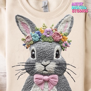 May include: A gray embroidered bunny wearing a pink bow and a flower crown with pink, blue, and green flowers. The bunny is on a beige sweatshirt with the text "ARTSY DIGITAL STUDIOS" in the top right corner.