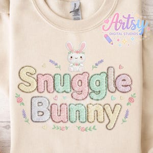 May include: A cream-coloured sweatshirt featuring the words "Snuggle Bunny" in pastel shades. The design includes a bunny wearing a floral crown, with floral and heart accents. The sweatshirt has a crew neck.