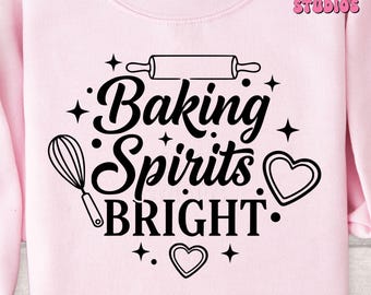 May include: A light pink sweatshirt with the words "Baking Spirits Bright" in a black script font. The design includes a rolling pin, whisk, and heart-shaped cookie cutter. The brand name "Artsy Digital Studios" is in the upper right corner.