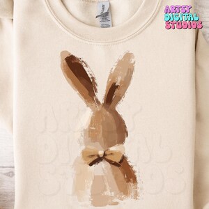 May include: A beige sweatshirt with a brown painted bunny wearing a bow tie. The bunny is facing away from the viewer. The text "ARTSY DIGITAL STUDIOS" is printed in the top right corner of the sweatshirt.