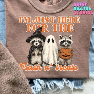 Peut inclure: T-shirt mauve avec l'inscription "I'M JUST HERE FOR THE Trash n' treats". Le graphisme représente un raton laveur avec un chapeau de sorcière, un fantôme et un raton laveur tenant une citrouille. Marque Comfort Colors.