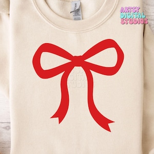 May include: Cream-colored sweatshirt featuring a large, bright red bow graphic. The bow is centered on the chest and has long, flowing ribbons. The text "Artsy Digital Studios" is in the upper right corner.