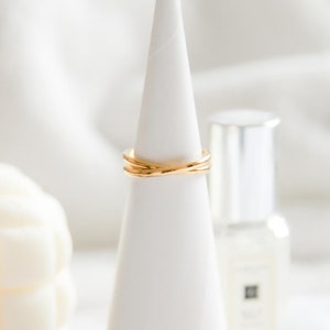 May include: A gold ring with a simple, elegant design. The ring is made of three bands that are intertwined. The ring is displayed on a white cone-shaped stand.